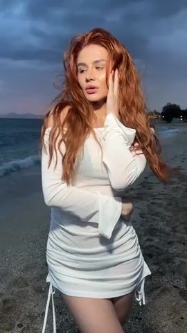 A red-haired woman in a white dress stands on a beach. The sky is cloudy and the sea is visible behind her. She crosses her arms and looks around.