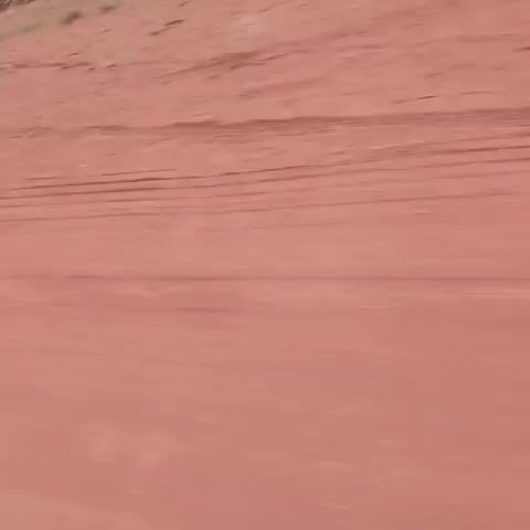 The video shows a reddish-brown dirt road from a first-person perspective, likely from a moving vehicle. Tire tracks are visible in the dirt. Green vegetation lines the edge of the road. The sky is overcast. The camera angle tilts upward at times, revealing more of the surrounding environment. A small portion of a person's orange shirt or jacket can be seen in the lower part of the frame.