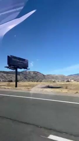 The video shows a view from a car moving on a highway on a sunny day. The road is cracked and has white lane markings. A sign indicates a closure ahead when flashing. A billboard advertising "More than you imagine" is visible, with mountains in the background. The sky is clear and blue. There are hints of reflections within the windshield of the car.