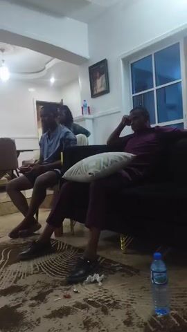 Here is a description of the video:

The video begins inside a house. Two men are sitting on a couch in the living room, and a water bottle is on the floor nearby. Then, the camera pans to a girl carrying a baby on her back. She is wearing a purple shirt that says “Milano de Rouge”, black shorts, and dark blue shoes. She walks around the room, and there is shelving and a mirror in the background.