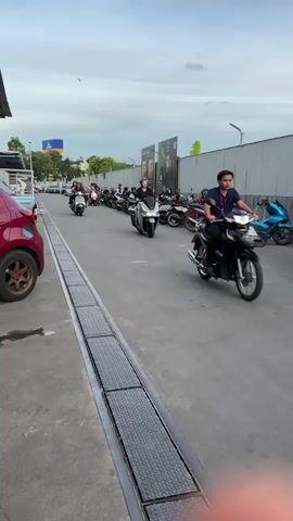 The video shows a street scene with several motorcycles parked along a wall. A person is riding a silver scooter in the foreground, with another person on the back. In the background, several other people are riding motorcycles and scooters. The sky is cloudy, and there is a building with signs in the background. The video is shot from a moving vehicle, giving a blurred perspective of the scene.