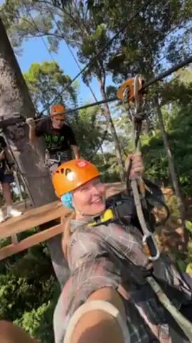A woman is riding a zip line through a forest. She is wearing an orange helmet and safety harness, and she is holding onto the zip line mechanism with one hand. The woman is smiling broadly and appears to be enjoying the experience. The zip line runs through a dense forest with tall trees and lush green foliage. Another person is seen on a platform at the beginning of the zip line course. The sky is clear and blue. The camera appears to be mounted on her wrist, providing a first-person view of the ride.