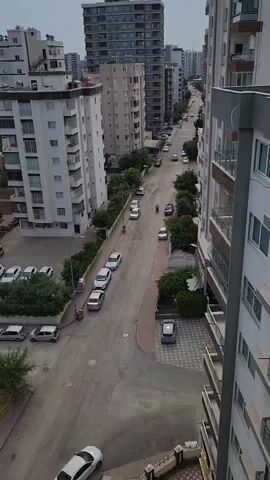 The video shows an aerial view of a street lined with tall residential buildings and parked cars. The street is paved, and there are trees and sidewalks along the sides. Several cars are driving on the street, and a motorcycle is also visible. The sky is overcast, suggesting a cloudy day. The overall impression is that of a typical urban street scene.