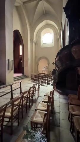 The video pans across the interior of a church. Rows of empty wooden chairs with woven seats are arranged in the nave. The ceiling features pointed arches and ribbed vaults. Natural light streams in from a large arched window at the far end of the church, illuminating a stained-glass window above the altar. A pipe organ is visible on a raised platform behind the altar. The floor is paved with stone tiles. The overall impression is one of quiet solemnity and architectural grandeur.