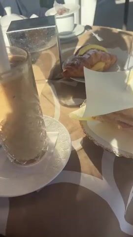 Here is a description of the video:
The video begins with a close-up of a table outside a cafe. On the table, there is a tall glass of iced coffee with a straw, a croissant, a sandwich, a cup, and a metal napkin holder. A man is visible in the background talking on a cell phone. The camera then moves up to show a woman's face momentarily, with bright sunlight behind her. The camera then moves back to show the table again, with the man still on his phone. Finally, the camera pans up to show the cafe umbrella, the street, and the view of the water and mountains in the distance on a sunny day.