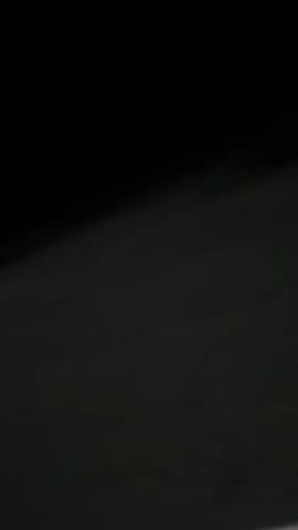 The video is taken from inside a vehicle at night. The camera is angled downwards and to the left, showing the interior of the car, specifically the dashboard area. The vehicle appears to be moving as the streetlights and trees blur in the background. The road is visible, with visible cracks in the asphalt. There is a streetlamp illuminating the scene. A triangular warning sign is visible in the distance, illuminated by the car's headlights. The overall impression is of a night drive on an empty road.