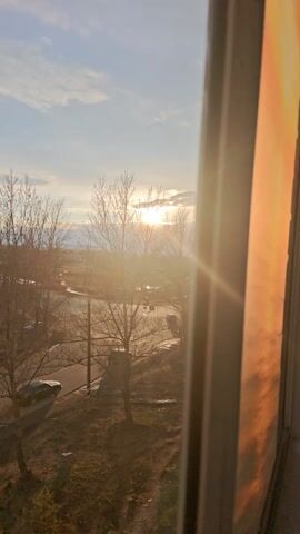 The video starts with a view through a window. Outside, the sun is visible through bare trees, and a car is driving down a road. The camera then pans to the right to focus on an image affixed to the window. The image is a bright orange and yellow scene resembling a sunrise or sunset over water. The camera focuses primarily on the window image.