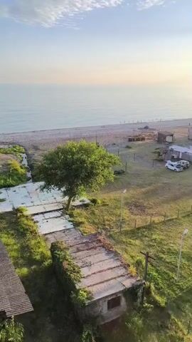 This is an aerial drone footage of a coastal area at sunset. In the foreground, there's an overgrown abandoned structure with vines covering its walls and roof. Next to it, a path leads towards a grassy field. The field has a playground with swings and climbing structures. Several people are scattered across the field, some appearing to be playing a game, possibly soccer. Further back, there are a few cars parked near some buildings. Beyond the field, the calm sea stretches to the horizon, reflecting the warm hues of the setting sun, which creates a bright glow in the sky. Buildings and trees dot the coastline.