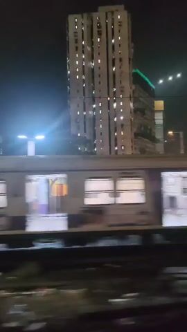 This is a shaky, handheld video taken at night from a moving vehicle. The video shows a train stopped at a station with a tall, illuminated building in the background. The train cars are white with purple and yellow stripes. The building appears to be an apartment complex with many windows lit from within. There are streetlights and possibly other lights in the distance. The area around the tracks appears to be dark and somewhat littered. The video captures the train and the building in motion, suggesting the vehicle is traveling alongside the train.