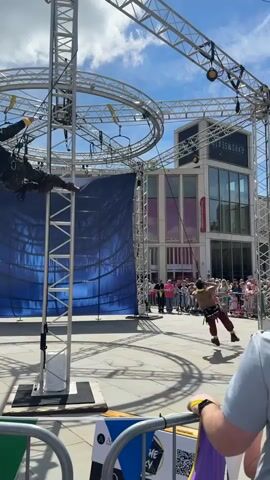 Here's a detailed description of the video:

The video shows a street performance in a public square.  A metal structure stands in the center, consisting of a vertical truss tower and a large circular truss suspended above. A blue sheet hangs behind the structure, possibly to hide equipment or create a backdrop. 

Several performers dressed in costume are present.  One individual is suspended upside down from the circular truss, held by ropes. Others on the ground enact a fight scene, including punches, shoves, and aggressive movements.  

A crowd of spectators watches from behind a metal barricade.  The "Glassworks" building is visible in the background, suggesting an urban setting.

The camera is held at ground level, providing a clear view of the performance and surrounding environment.  The weather appears sunny and bright.