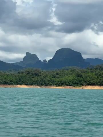 The video shows a body of turquoise water in the foreground. The water is calm with small ripples. In the middle ground, there is a shoreline covered in green vegetation. Behind the vegetation, there are large, dark green mountains under a cloudy sky. The clouds are grey and white, with some appearing dark and heavy. The overall scene is a natural landscape.