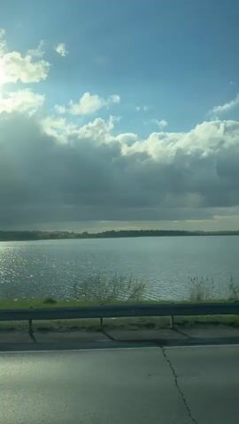 The video shows a view from a moving car, looking out over a lake on a partly cloudy day. The water is slightly rippled and reflects the sunlight. A safety barrier runs along the side of the road, with some vegetation growing between it and the water. In the distance, trees line the far shore of the lake. The road surface is visible in the foreground.