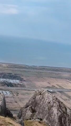 A drone shot captures a panoramic view of a vast, arid landscape under a hazy sky. The foreground is dominated by a large, jagged rock formation bathed in sunlight, with dry grass and sparse vegetation clinging to its slopes. Beyond the rock, the terrain unfolds into a patchwork of cultivated fields, some appearing fallow and others showing signs of agricultural activity. A winding road or highway cuts through the valley, with what appear to be industrial or agricultural buildings, possibly greenhouses, visible in the middle ground. In the distance, a body of water, likely a sea or a large lake, stretches towards the horizon, its surface rippling under the diffuse light. The overall impression is one of a remote, sparsely populated, and dry environment.