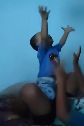 A child in a blue t-shirt with a cartoon bear print is sitting on an adult's lap. The child's arms are raised as they play with a red balloon. The adult is holding the child and the balloon. The scene appears to be indoors, with a light blue wall in the background and a patterned blanket beneath them.