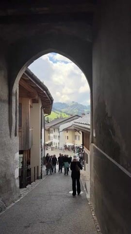 The video shows a view through a stone archway onto a cobblestone street in a European village. People are walking down the street towards a small town square in the distance. Buildings line both sides of the street, with wood and stucco exteriors and tiled roofs. Beyond the buildings and street is a view of rolling green hills under a blue sky with puffy white clouds. A street sign is visible on the right side of the arch.