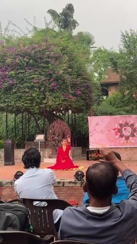 A young woman performs a dance on a brick stage in an outdoor setting, in front of an audience of a few people, some of whom are filming her with their phones. She wears a red dress with gold trim and jewelry, and she's sitting on the floor with her hands raised. Behind her is an intricate metal sculpture of a deity, and a tree with purple flowers. There's also a pink banner to the right of the sculpture with an image of a lotus flower on it. Someone walks behind the dancer. The setting appears to be a garden or courtyard, surrounded by greenery.