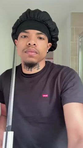 A young man wearing a black durag and a black t-shirt stands in a bathroom, holding a sword. The sword has a silver-colored blade and a white handle. He is wearing a silver ring on his left hand. The man looks directly at the camera, raising the sword over his right shoulder and then lowering it and examining the handle. The bathroom has white walls and a shower behind him. The door to the bathroom is open, revealing another room in the background.