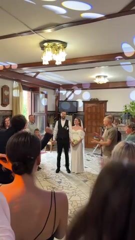 A wedding celebration is taking place in a well-lit indoor venue. A bride and groom stand center frame, facing the camera. The bride wears a white wedding dress with off-the-shoulder sleeves and a veil. The groom is dressed in a black vest, white shirt, and black pants. They are holding hands and smiling. Around them, a crowd of guests are clapping and cheering. Some guests are visible in the foreground, with their backs to the camera, and others are in the background. The room has wooden accents on the ceiling and walls, and decorative lighting fixtures. There is a television screen visible in the background. The floor has a mosaic tile pattern. The overall atmosphere appears festive and joyful.