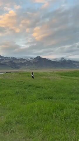The video captures a serene, wide-angle view of a person running across a vast green field in Búðir, Iceland. The individual, dressed in dark athletic wear, is jogging from left to right in the middle ground, appearing small against the expansive landscape.

In the foreground and midground, lush green grass stretches across the frame, indicating a fertile plain. Beyond the field, a majestic range of dark, rugged mountains dominates the background. Several peaks are visible, some partially covered with snow or shrouded in low-hanging clouds, adding to the dramatic scenery. A body of water, possibly a river or an inlet, can be seen winding through the landscape on the left side, reflecting the subdued light.

The sky is partly cloudy, displaying a beautiful mix of soft grey, white, and subtle hints of orange and pink, particularly on the underside of the clouds. This warm lighting suggests either an early morning sunrise or a late evening sunset, casting a gentle glow over the scene. The overall atmosphere is one of tranquility and natural grandeur, with the runner enjoying the vast, open space of the Icelandic wilderness. No visible text is present.