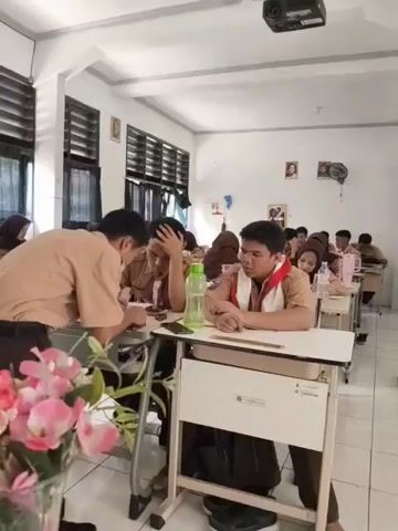 The video shows a classroom scene where two male students are interacting. One student, wearing a brown uniform with red and white insignia on the collar, is leaning over a desk, seemingly talking to another student sitting at the desk. This seated student, also in a brown uniform with similar insignia, has his hand on his forehead and appears to be looking down or in thought. A green water bottle is visible on the desk between them. In the background, other students are seated at desks, and the room contains a projector mounted on the ceiling, windows with blinds, and decorative elements like flowers in a vase in the foreground. The overall impression is of a school setting during a lesson or activity.