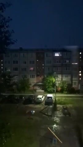 The video shows a dark, nighttime scene of a residential building. The building is several stories tall with multiple windows, some of which are lit from within. In front of the building, there is a small playground with a seesaw and some other equipment. There are also several cars parked in the foreground. The ground appears to be wet, suggesting it might have rained recently. The overall atmosphere is quiet and still.