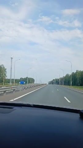 The video shows a view from inside a car, looking out at a highway. The road is empty and stretches into the distance. There are trees lining the sides of the road and streetlights. The sky is blue with some clouds. The dashboard of the car is visible in the foreground.