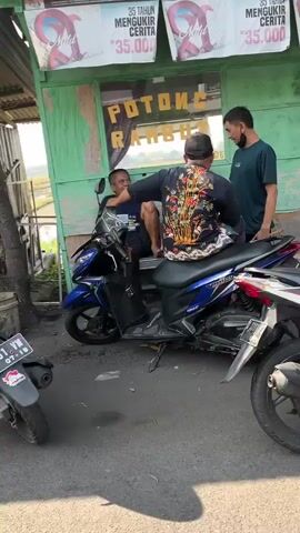 The video shows three men in an outdoor setting, likely a street or sidewalk, in front of a green building with signs advertising haircuts. A blue and black scooter is the central object, with two men sitting on it and one man standing. The man in the foreground, wearing a black shirt with an ornate, colorful design on the back, is seated behind the rider of the scooter, facing away from the camera. His left arm is extended, and he appears to be holding or gesturing towards the rider. The rider of the scooter, wearing a dark blue t-shirt and jeans, is facing forward and appears to be talking to the standing man. The standing man, wearing a dark blue t-shirt and black pants, is facing the rider and the man behind him, his right arm is raised, and he seems to be engaged in a conversation or interaction. The environment suggests a commercial area with parked motorcycles and a simple structure. Sunlight indicates it is daytime.