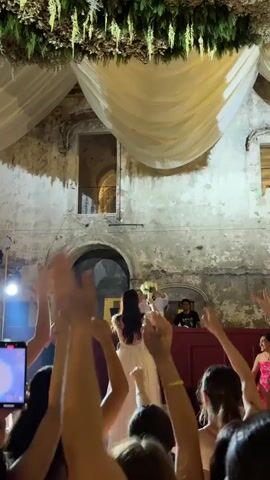The video shows a wedding party happening in what looks like the courtyard of an old building. There’s a bride being held up by a man, presumably the groom, throwing her bouquet into a crowd of outstretched hands. A DJ is visible behind them on a raised platform with his headphones on, and another man wearing a t-shirt is standing next to him. There are hanging decorations and drapery above. The crowd seems excited, with many people holding up phones to record the moment. The architecture of the building features stone walls and archways.
