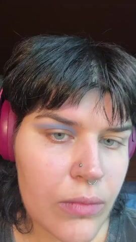 A person with dark hair and blue eyes is applying purple eyeshadow to their left eyelid with a neon green and pink makeup brush. The person is wearing pink headphones on their left ear and has a nose ring. The background is blurry but appears to be an indoor setting.