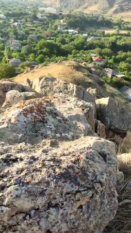 The video begins with a close-up of large, rough rocks in the foreground. The camera then pans up and to the right, revealing a valley with a village nestled among trees. Houses with red and grey roofs are visible. Beyond the village, a road winds through a green landscape, which gradually transitions into rolling hills and mountains in the background. The sky is clear and blue. The overall scene is a panoramic view of a rural village surrounded by natural landscapes. The camera movement is smooth and deliberate, allowing for a comprehensive view of the environment. The lighting suggests it is daytime, likely late afternoon or early morning, given the warm tones and shadows. There are no people or animals visible in the video. The focus is solely on the landscape and the settlement within it. The video ends with a slight shake, indicating the end of the recording.
