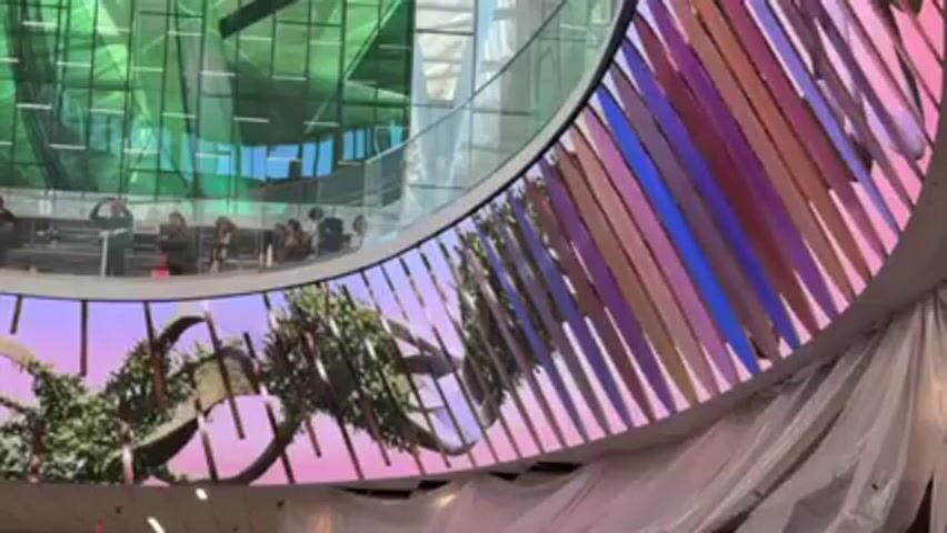 The video shows a large, curved LED screen displaying a nature-themed animation with green vegetation against a pink and blue sky backdrop. The screen is segmented with vertical bars. Above the screen is a balcony area with people looking down, some taking pictures. The building has green-tinted windows. There's some white material draped around the bottom of the screen. The camera slowly zooms in on the display.