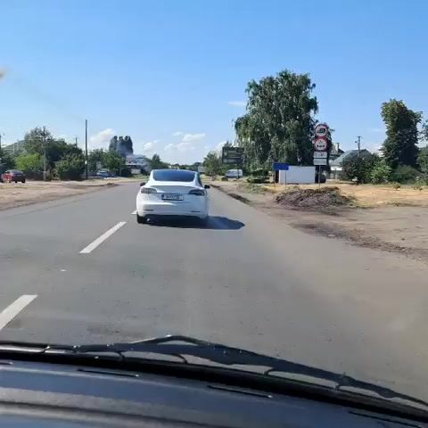 The video shows a view from inside a car driving on a paved road. A white Tesla is in front of the car, in the same lane. The road has lane markings. There are trees and some buildings on the right side of the road, as well as a billboard. The sky is blue with some clouds. Another car, a silver SUV, is in the left lane. The Tesla briefly emits a white puff, possibly smoke or steam, from its rear. The video continues for a few seconds with the cars moving forward.