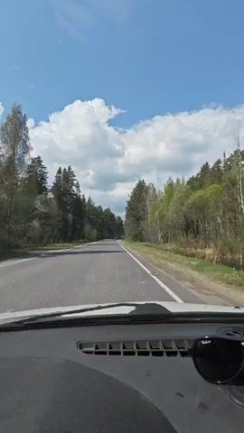 The video shows a view from inside a vehicle traveling along a straight, paved road. The sky is blue with scattered white clouds.  On either side of the road is dense forest. Sunglasses rest on the dashboard in the foreground. The camera appears steady, and the road is clear.