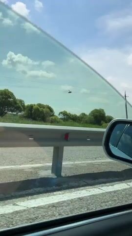 The video shows a view from inside a moving vehicle looking out a side window. The car is traveling on a road with a metal guardrail visible on the left. Beyond the guardrail are green fields and scattered trees under a bright blue sky with white clouds. Telephone poles are also visible in the distance. The sun is shining, and the overall scene suggests a rural or semi-rural environment during the daytime. The video appears to be a short clip, likely capturing a moment of travel.