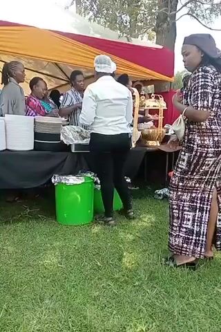 The video shows an outdoor event with a food serving station set up under a tent with red and yellow fabric. Several people are gathered around a table laden with food and serving dishes. To the left, stacks of white plates and bowls are visible. A woman wearing a white shirt and black pants, with her hair covered by a hairnet, is serving food from a foil-covered dish. Other individuals are seen standing or walking around the serving area. Two green cylindrical bins with silver lids are placed on the ground in front of the table. The ground is covered in green grass. In the foreground, a woman in a colorful patterned dress is walking away from the camera. Another woman in a black and white patterned dress is partially visible on the right side of the frame. The overall scene depicts a catered event with a buffet-style food service.