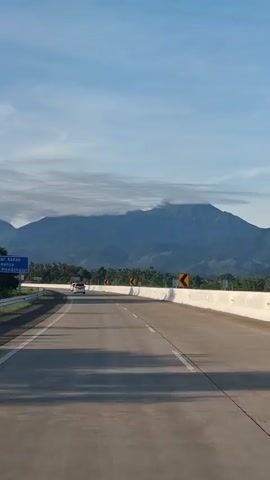 A view from a moving vehicle on a highway shows a scenic mountain landscape under a clear blue sky with scattered clouds. The highway has two lanes in the direction of travel and is bordered by white concrete barriers. Several cars are visible on the road ahead. Yellow curve warning signs are placed along the right side of the highway. The mountains in the background are covered in vegetation.