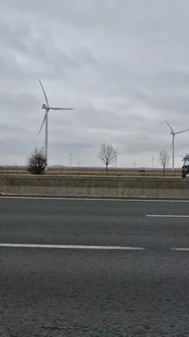 The video shows a highway with white lines marking the lanes. On the right side of the road is a concrete barrier with green grass and trees behind it. Wind turbines are visible in the distance. First, a truck with "XPOLogistics" printed on its side drives by. The camera continues moving forward, and several more wind turbines come into view. A triangular road sign and a small white car also appear in the background. The sky is overcast.