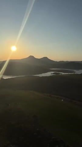 The video captures a serene sunrise over a vast landscape, likely near Yeşilhisar, Turkey. The bright sun, low on the horizon, casts a golden glow and long rays across the scene. In the foreground, green fields undulate across the terrain, leading towards rolling hills and mountains in the distance. A winding body of water, possibly a lake or reservoir, is nestled amongst the hills. As the video progresses, hot air balloons become visible, gracefully ascending into the clear morning sky above the hilly landscape, with the distinct sound of a burner audible. The scene evokes a sense of tranquility and awe, showcasing the beauty of the natural environment during an early morning hot air balloon flight.