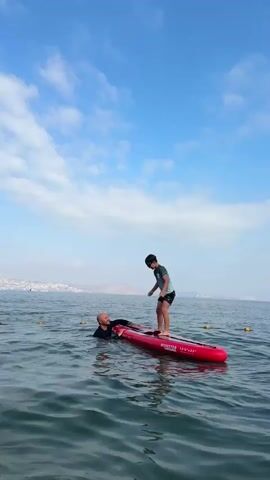 A man in a black shirt and shorts is in the water near a red inflatable stand-up paddleboard. He is holding onto the board. A boy in a blue and black swimsuit is standing on the paddleboard, facing the man. The boy appears to be in the process of jumping or falling off the board. The water is choppy, and the sky is blue with white clouds. In the distance, there is a coastline with buildings. The man is partially submerged in the water and is looking towards the boy. The boy's right arm is raised. The board has "MONSTER" written on it.