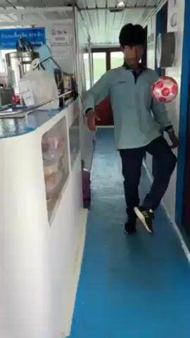 Here is a description of the video. 
A young man is seen in a narrow corridor of a boat, skillfully juggling a red can with his feet, seemingly without dropping it. The corridor has a blue floor, white walls and ceiling, and a counter to the left. He's dressed in a light blue long-sleeved shirt and dark pants. After the can drops, he looks up at the camera as another person approaches.