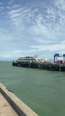A ferryboat is docked at a pier under a partly cloudy sky. The water is a greenish hue. The ferry is white with a prominent superstructure and a large mast. It appears to be a passenger ferry. The pier is constructed of concrete pilings and a wooden deck. There are also some blue structures and red coiled ropes on the pier to the right of the ferry. The foreground shows a concrete walkway. The overall scene is calm and suggests a functional port.