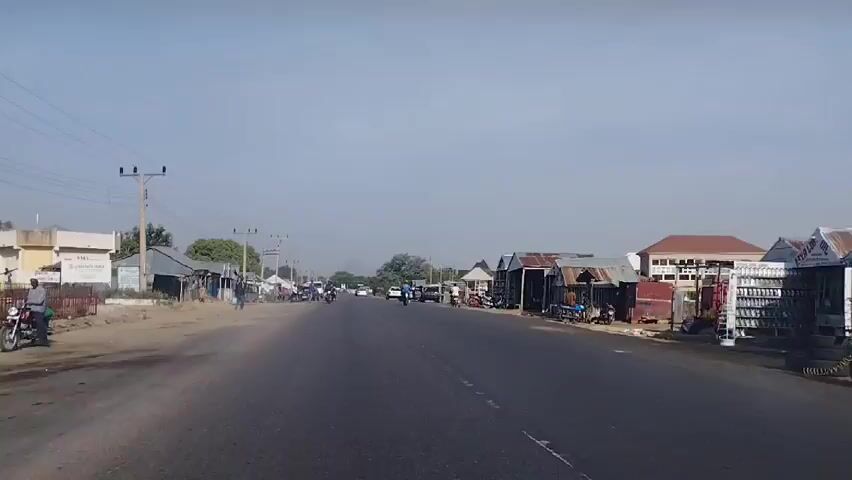 The video shows a view from a moving vehicle traveling along a paved road that bisects a rural town or village. The sky is overcast, providing a muted, diffused light. The road surface is generally well-maintained, with visible lane markings or dividers. 

On either side of the road, there are various structures ranging from commercial buildings and shops to residences. These buildings appear to be of modest construction, some with corrugated metal roofs. Several individuals are visible: some are standing by the roadside, others are walking, and several are riding motorbikes. Vehicles, including cars and motorcycles, are present, both parked and in motion. 

A prominent feature of the environment is the presence of utility poles with power lines running along the side of the road. The surroundings consist of a mix of natural and man-made elements, giving the impression of a semi-urbanized, developing area.