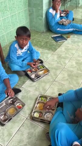 The video shows several young boys, dressed in blue and white school uniforms, sitting on a tiled floor and eating from metal trays. The room appears to be a common area or dining hall, with green tiled walls and a wooden stool in the background. The boys are interacting with each other and their food, with some smiling and others focused on their meals. In the background, two other children, one possibly a girl wearing a headscarf, are also eating. Another adult is visible in the distance, walking away from the camera. The trays contain various food items, including rice, vegetables, and what appear to be pieces of meat or fish. The overall atmosphere seems casual and communal, suggesting a school mealtime.