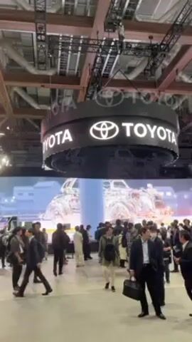 The scene takes place at a large indoor event, likely a trade show or conference, under high ceilings with exposed metal framework. A large, illuminated Toyota sign hangs from the ceiling. Below, a crowd of people walks through the event space, with most wearing masks. In the background, a large screen displays an abstract image of a vehicle. The overall atmosphere is busy.