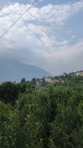 The video shows a scenic view of mountains and trees under a cloudy sky. In the foreground, there are lush green trees and bushes. In the middle ground, there is a cluster of houses on a hillside, surrounded by more greenery. In the background, majestic mountains rise up, partially obscured by clouds. A thin wire, possibly for electricity or communication, stretches diagonally across the frame from the top left. The overall impression is one of a peaceful, natural landscape.