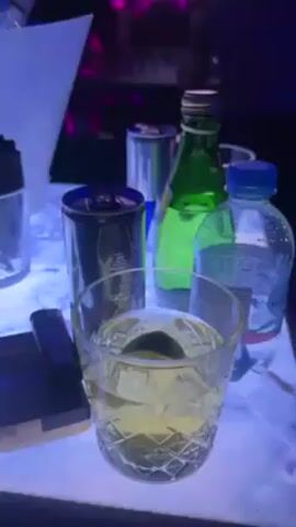 The video presents a close-up view of a bar table, lit with a vibrant blue light, with a variety of beverages and accessories. A cut-crystal glass filled with a light-colored liquid and ice sits prominently in the foreground. Adjacent to it is a dark-colored beverage can, a green glass bottle, and a plastic water bottle. A small, dark box with the word "SOBRAME" is visible on the left. Behind this collection, an ice bucket filled with multiple bottles, including a bottle of Ostoya Vodka, is visible. The background is blurry, suggesting a busy environment, with glimpses of people and ambient lighting indicating a nightclub or bar setting.