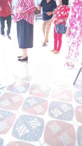 The video shows the legs and lower torsos of several people standing on a tiled floor. The tiles are square with a pattern of diamonds and squares in brown, beige, and grey. One person in the foreground is wearing a black skirt and black flats. Next to them, a person is wearing a red and white striped shirt and a black skirt. To the right, another person wears a red shirt with white polka dots and red pants. Further to the right, a person wears a pink and white tie-dye dress. In the background, the legs of two men are visible; one wears tan pants and black shoes, and the other wears dark pants and black shoes. The overall scene suggests an outdoor gathering or event.