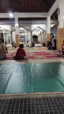 The video shows an indoor scene with several people sitting on the floor. The floor is covered with patterned red rugs in the background and a green tarp in the foreground. There are pillars supporting a tiled roof. In the background, there appear to be several tomb-like structures or altars. People are seated on the rugs facing these structures. Some individuals are wearing traditional Islamic attire, including kufi caps. The lighting suggests it is evening or indoors with artificial lighting. The overall atmosphere appears to be solemn or devotional.