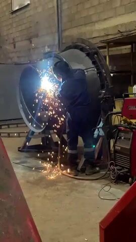 In a brightly lit industrial setting, a worker in a dark blue jumpsuit is welding a large, cylindrical metal structure.  The worker is facing the structure, with welding gear obscuring their face. Intense sparks and bright light emanate from the welding point. The cylinder is supported by a metal stand, and a welding machine sits nearby. The walls of the workspace are constructed of cinder blocks, with small windows near the ceiling. Foreground objects, including a red workbench, partially obscure the view. Wires and hoses are strewn across the concrete floor.