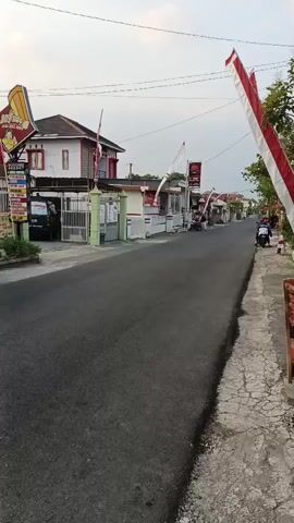 A black pickup truck drives down a paved road lined with buildings and trees. Red and white flags are visible on poles. The sky is overcast.